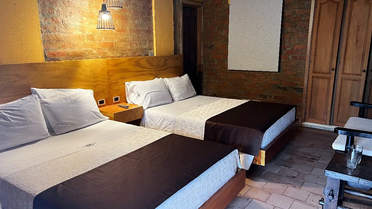 Habitaci&oacute;n Cu&aacute;druple: dos camas queen con cabeceros de madera y pared de ladrillo
